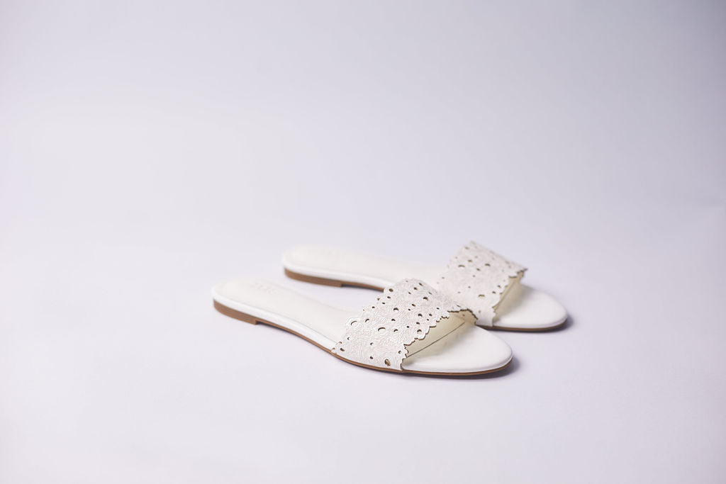 A pair of white leather slide sandals with laser-cut eyelet straps, displayed on a clean, light gray studio background.