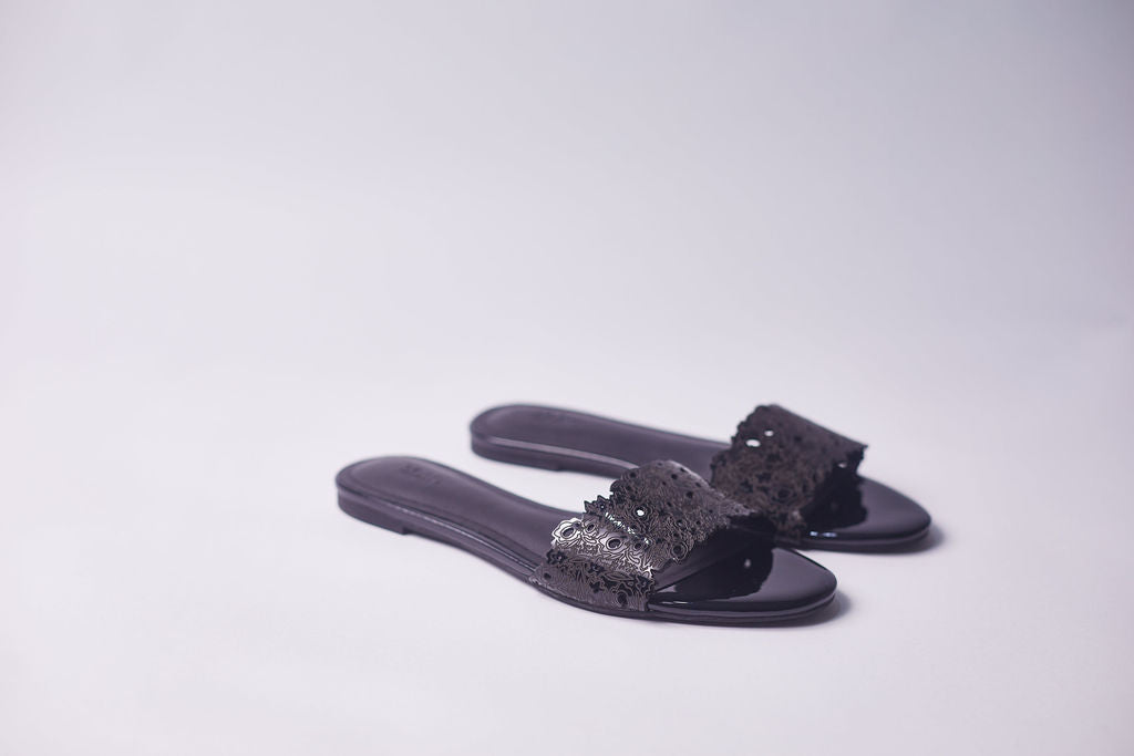 A pair of black slide sandals with textured, perforated straps, displayed on a clean light gray studio background.