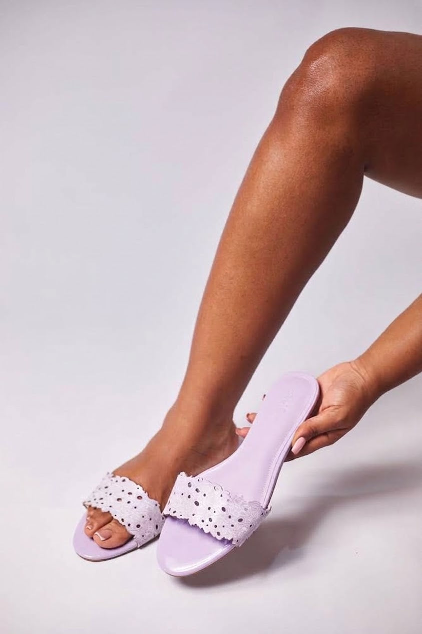 A person wearing lavender slide sandals with white lace-up straps and small black polka dots, with one hand adjusting the sandal on their foot against a light gray studio background.