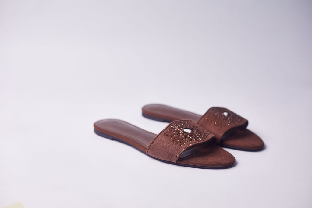 A pair of brown suede slide sandals with rhinestone-embellished straps, photographed on a plain light gray background.