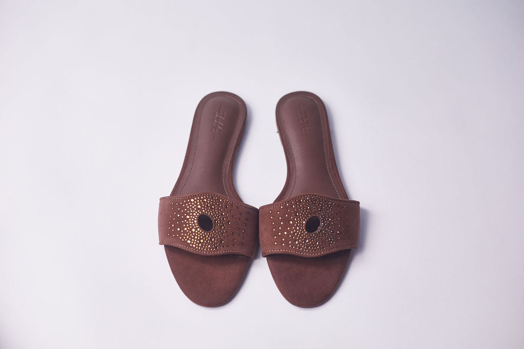 A pair of brown slide sandals with a wide strap featuring a circular cutout and a border of small metallic studs, shown on a light gray background.