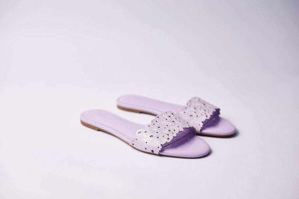 A pair of lavender flat slide sandals with a wide cutout strap, shown on a clean light gray studio background.