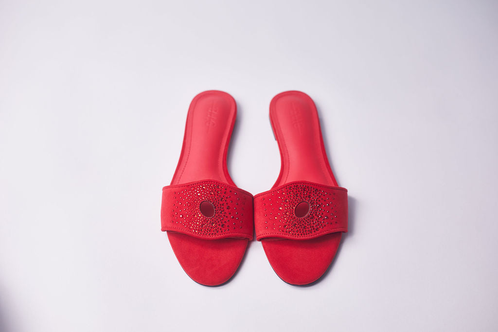A pair of red slide sandals with textured straps and circular cutouts, displayed on a light gray background.