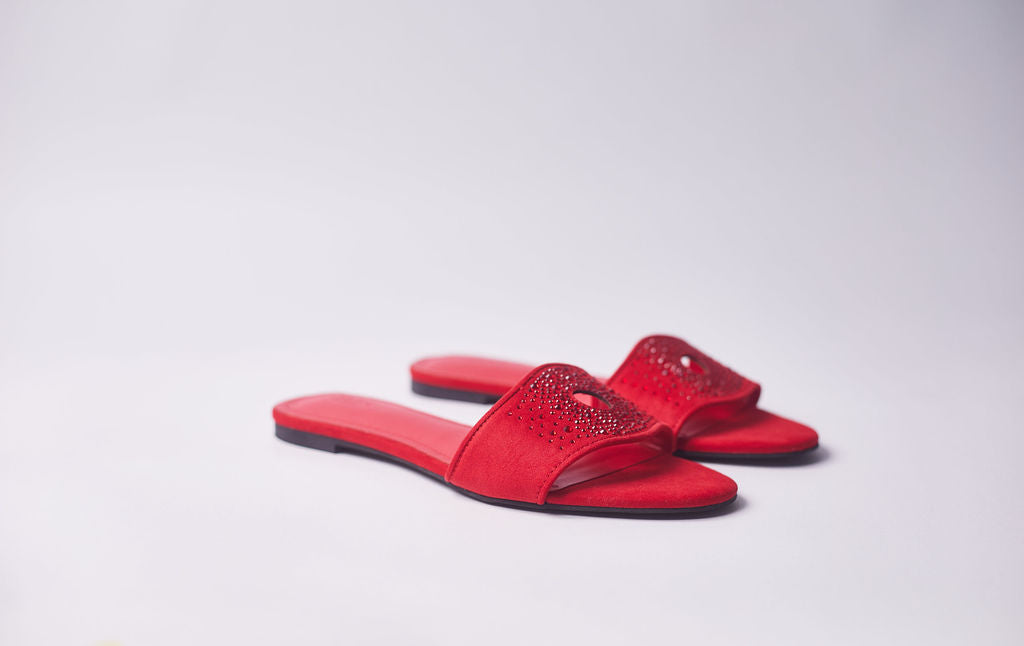 A pair of red slide sandals with open toes and rhinestone embellishments on the straps, shown on a light gray studio background.