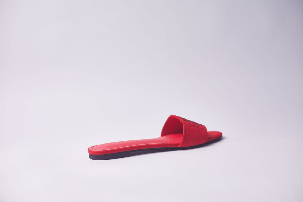 A single red slide sandal with a flat sole, photographed from a side profile against a clean, light gray studio background.