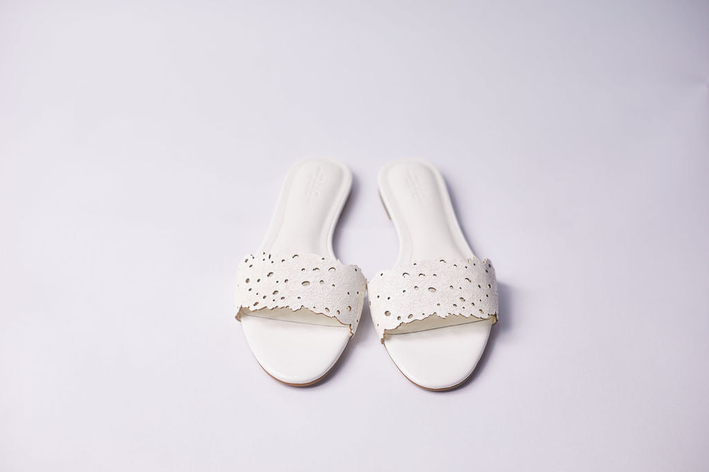 A pair of white slide sandals with laser-cut eyelet straps, displayed on a clean, light gray background.