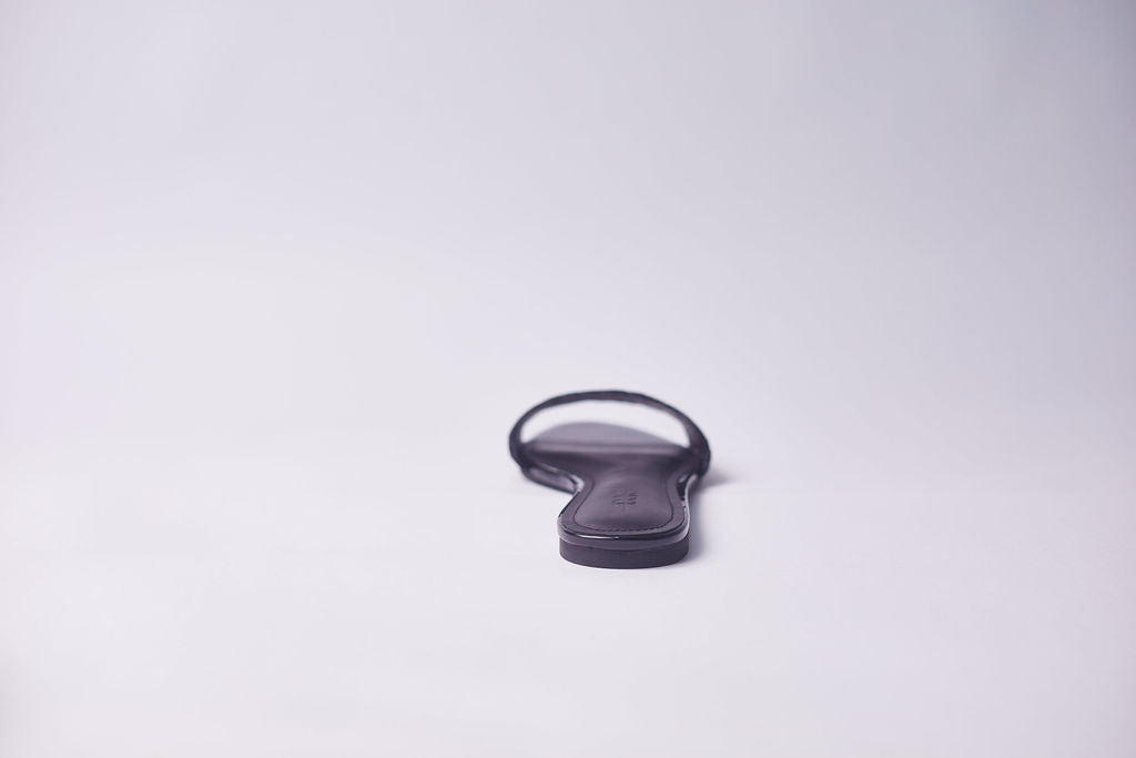 A single black flat sandal shown from the back on a seamless light gray studio background.
