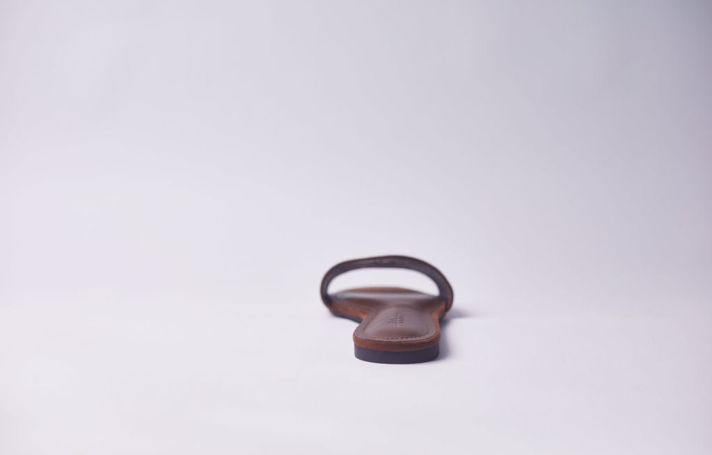 A single brown leather flat sandal shown from the back on a clean, light gray studio background.