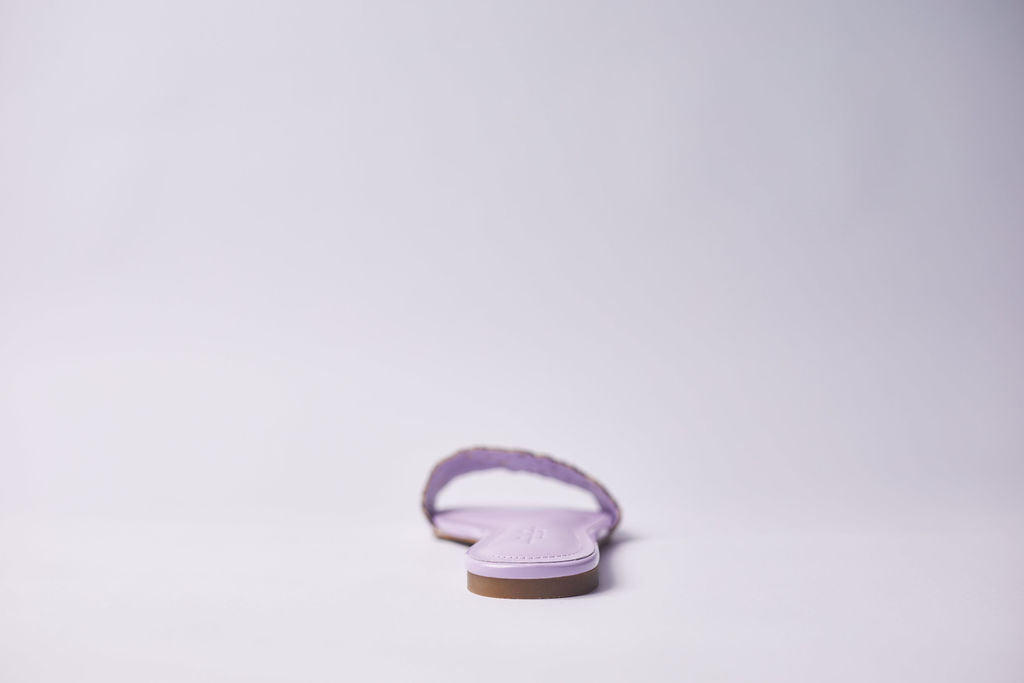 A single light purple flat sandal with a textured strap and a brown sole, photographed from the back against a clean white background.